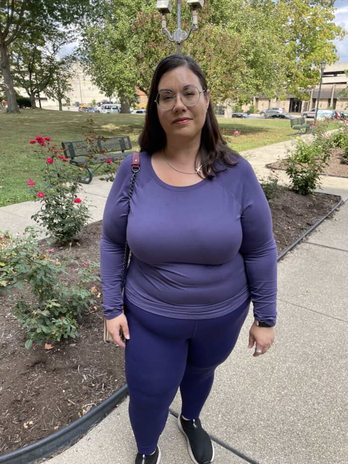 Woman with glasses in purple shirt with dark hair. Standing outside. A garden with red flowers can be seen behind her.