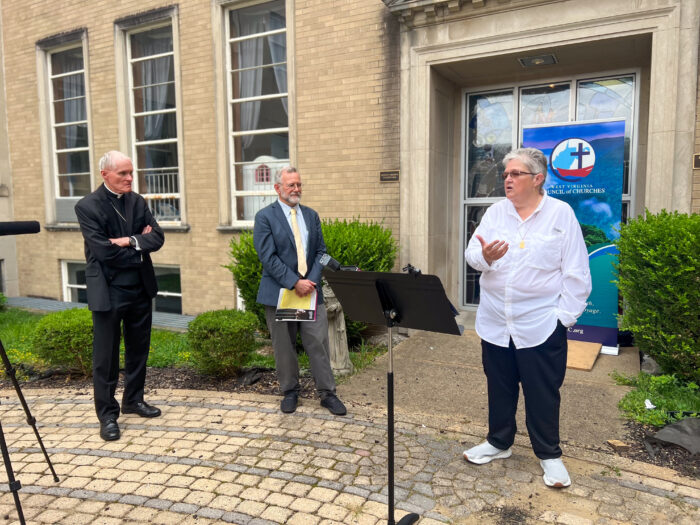 Three people stand in front of a building. One woman and two men. The woman has short white hair and is speaking to a group. She has a stand in front of her.